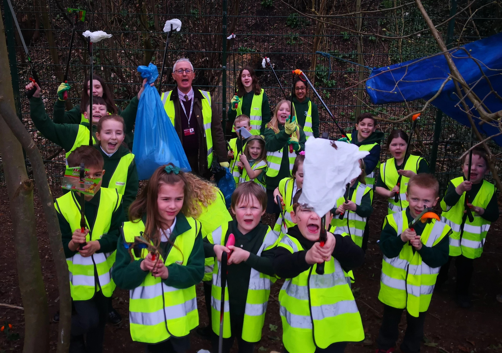 Group photo of school children wearing bright yellow high vis jackets holding litter pickers with rubbish up in the air.